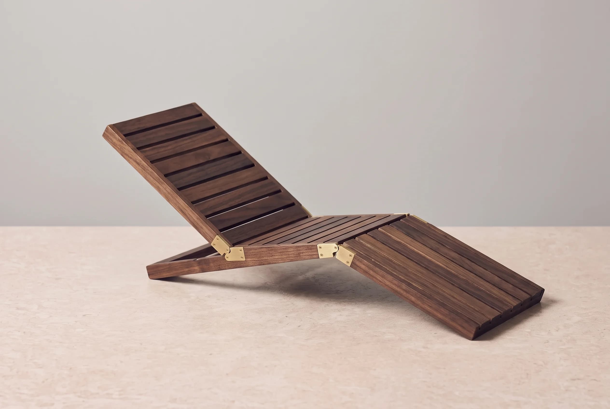A closed wooden sun lounger with brass hinges lies folded on a stone surface against a grey wall, lit from the side.