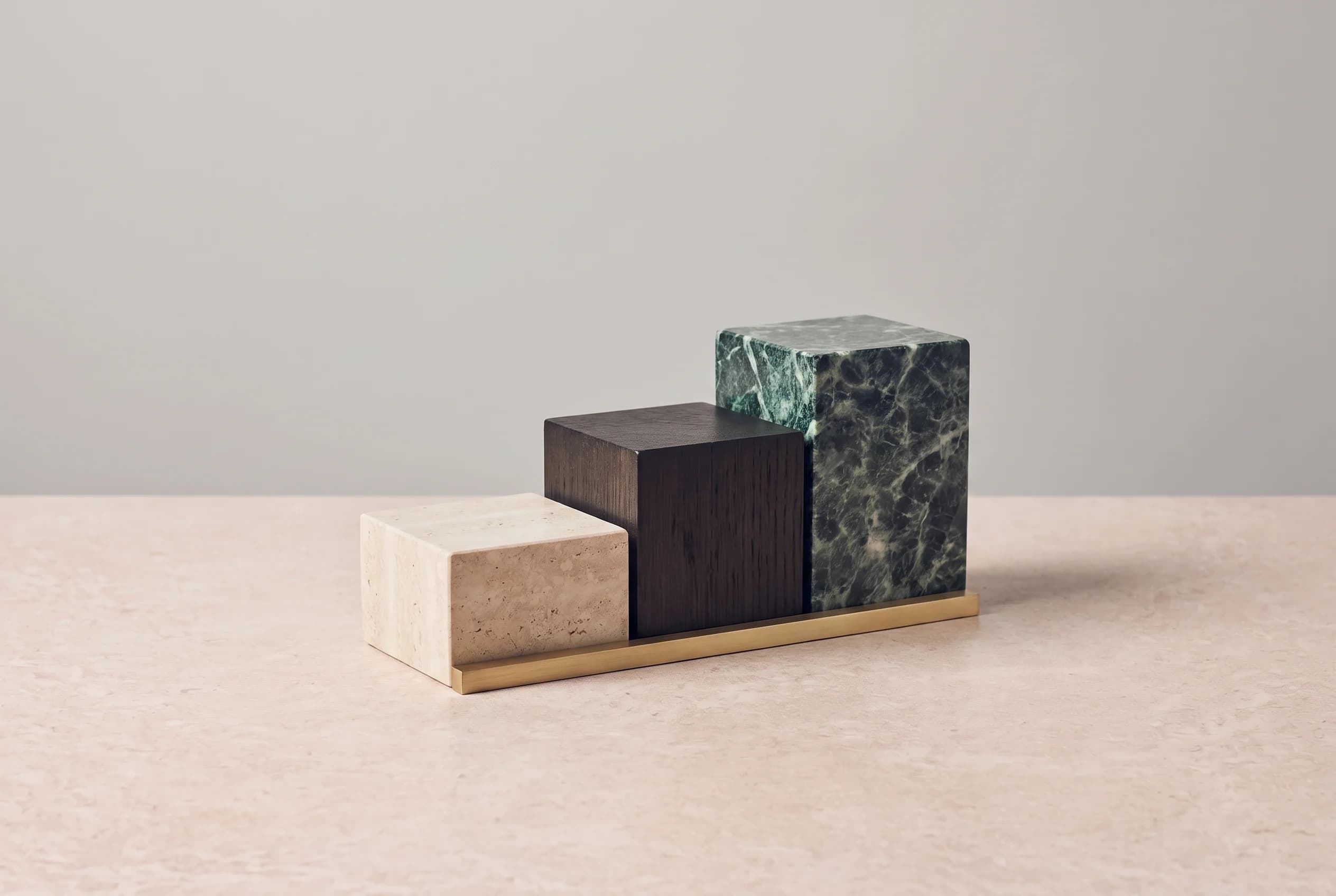 Three rectangular blocks of increasing height in travertine, dark oak and green marble stand in a row on a stone surface, with a thin brass bar at their base.