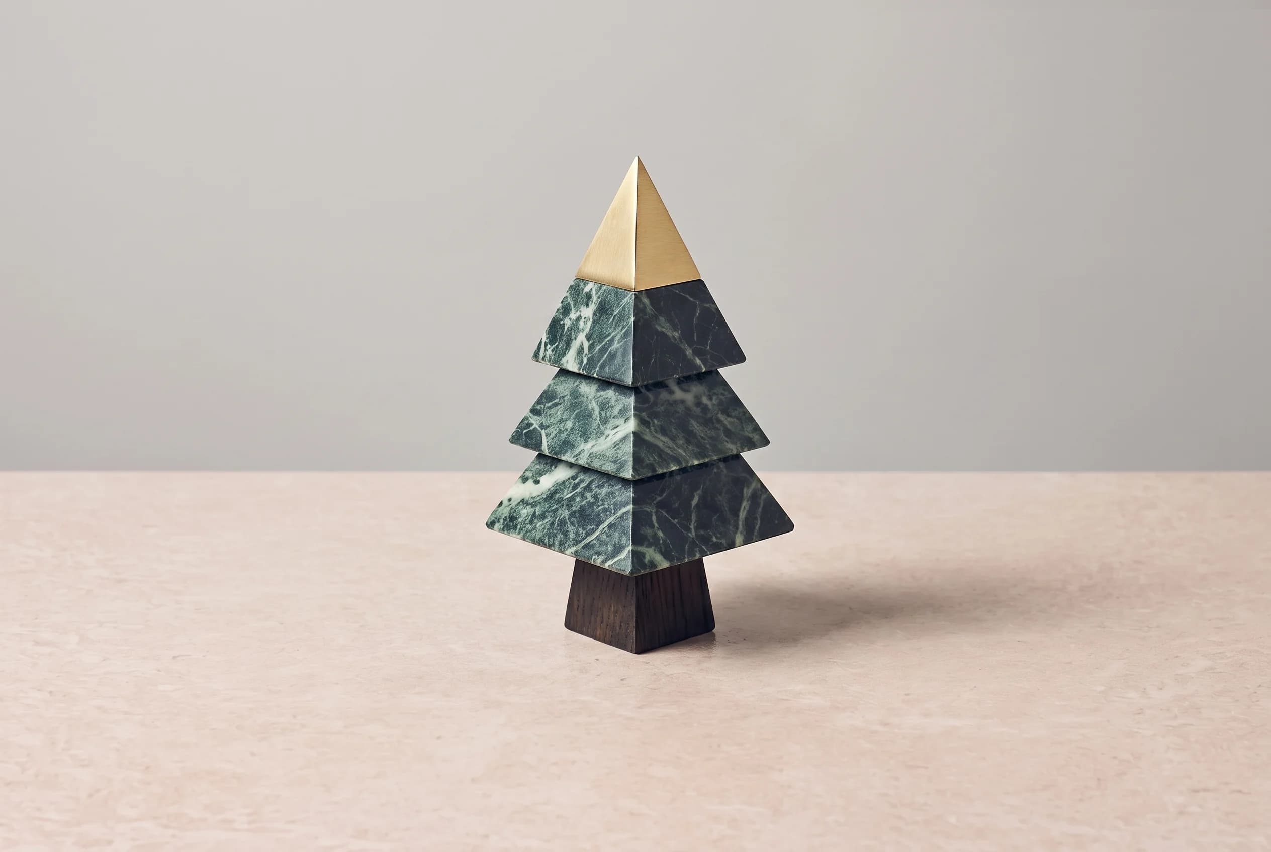 A geometric fir tree made of dark oak, green marble and brass stands on a stone table against a grey wall.