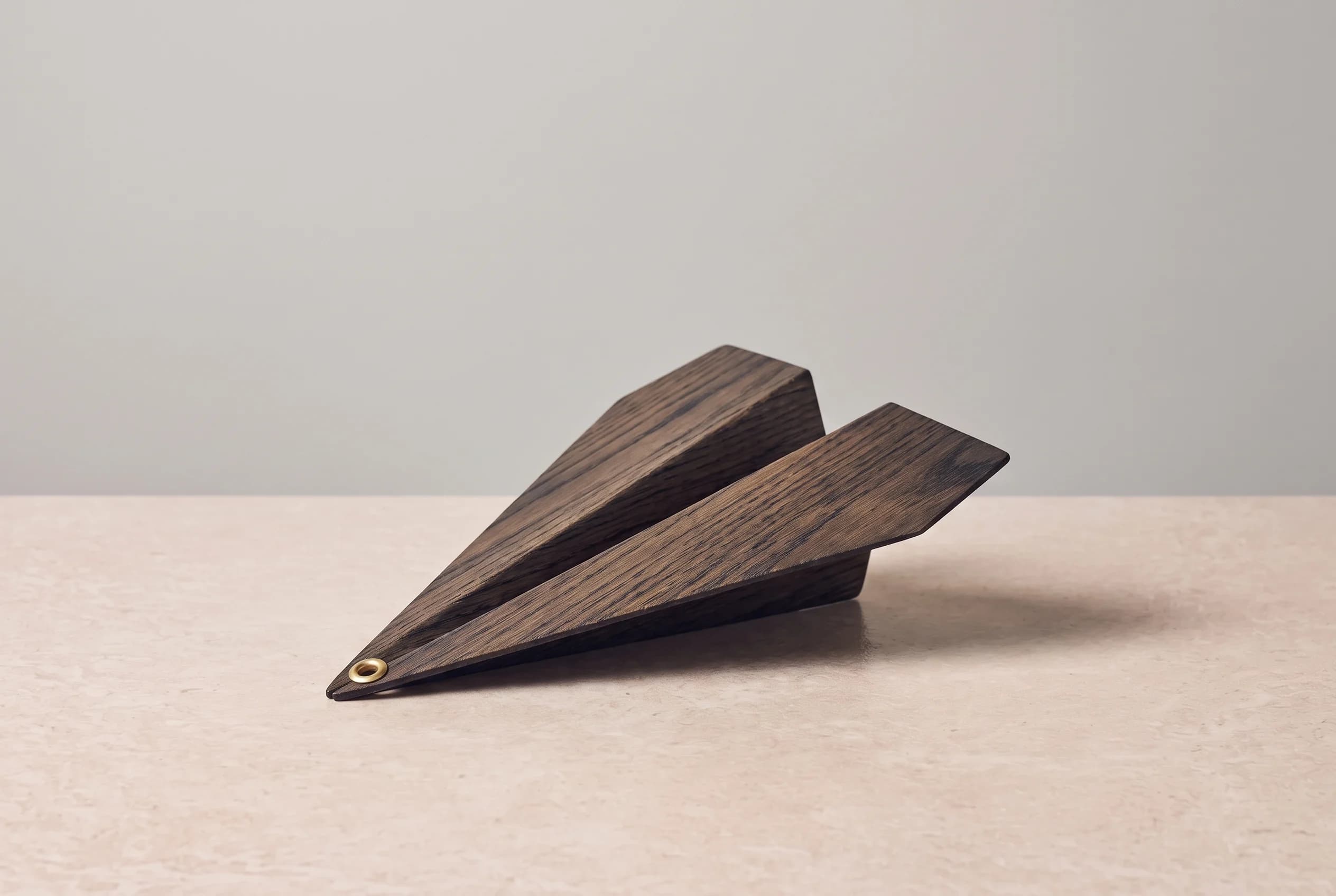 A folded triangular sail made of dark smoked oak with a brass grommet, lying on a travertine surface against a grey wall.