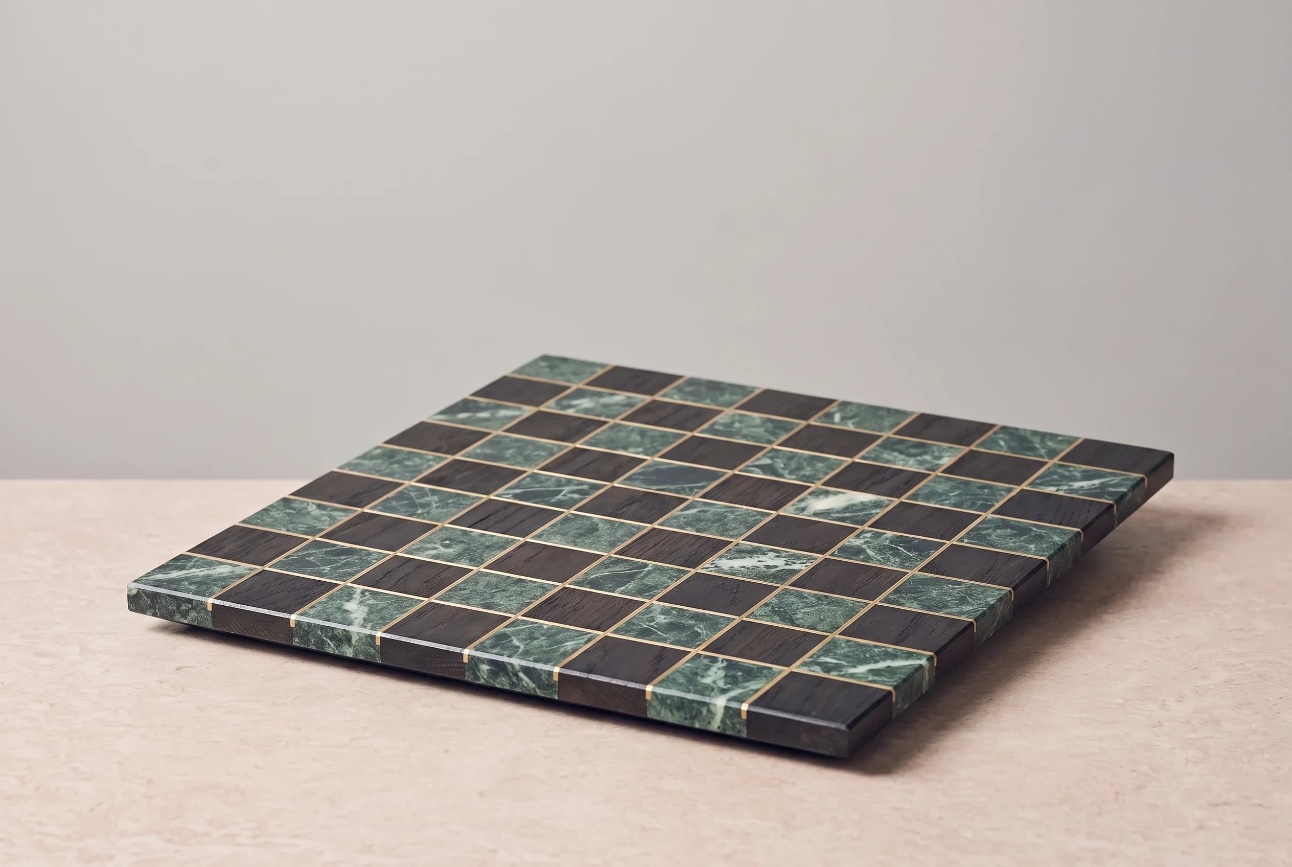 A still life of a square chessboard inlaid with Verde Alpi marble, dark smoked oak and brushed brass lines, resting on a beige stone table against a grey wall.