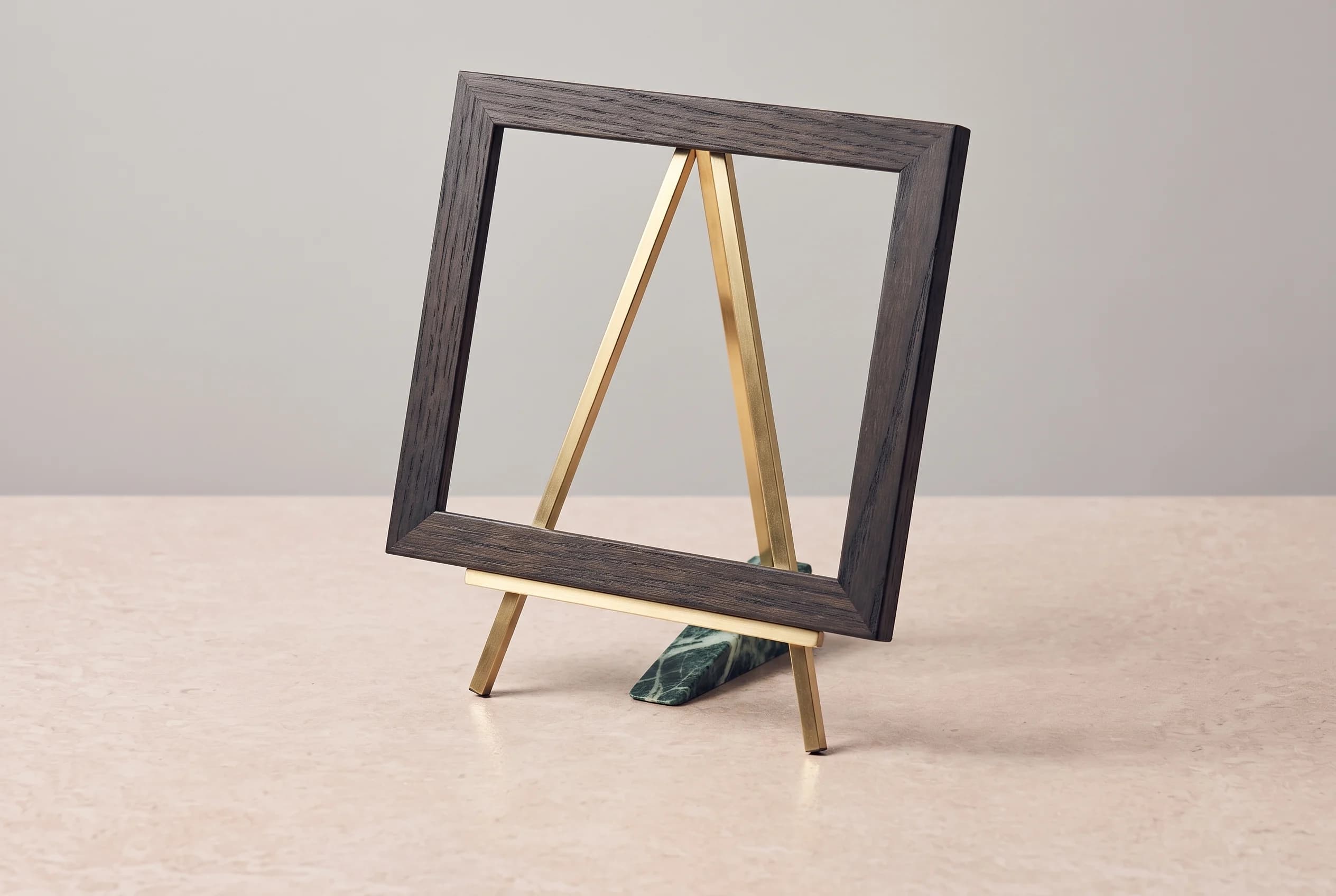 An empty dark oak picture frame rests on a brushed brass easel on a pale stone surface, lit from the side against a grey wall.