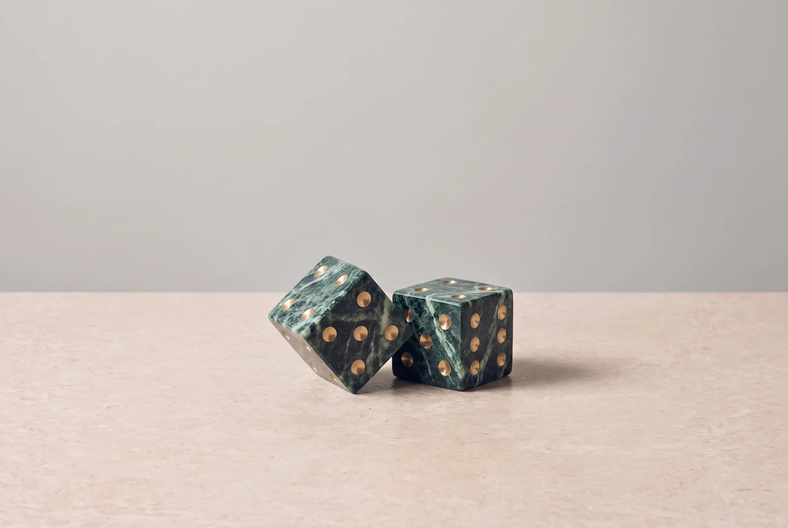 Two marble dice with brass inlaid pips sit at an angle on a travertine surface, cast in soft side light against a grey wall and dark oak panel.