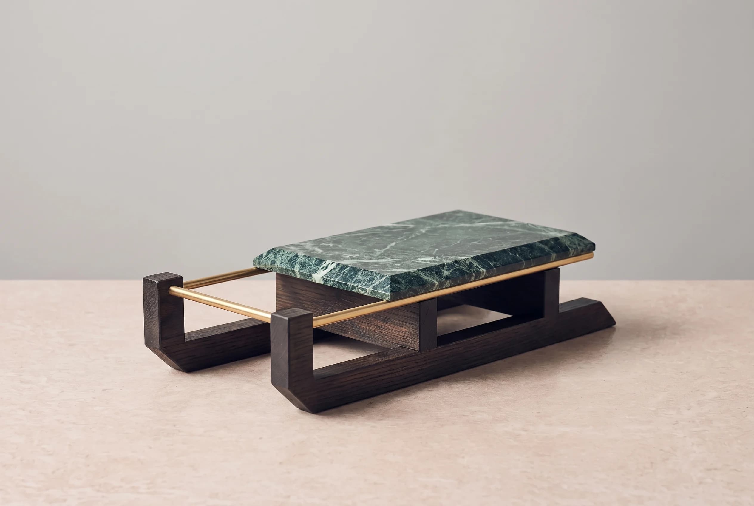 A still life of a wooden sledge with squared oak runners, a marble seat and a brass bar, standing on a stone surface against a grey wall.