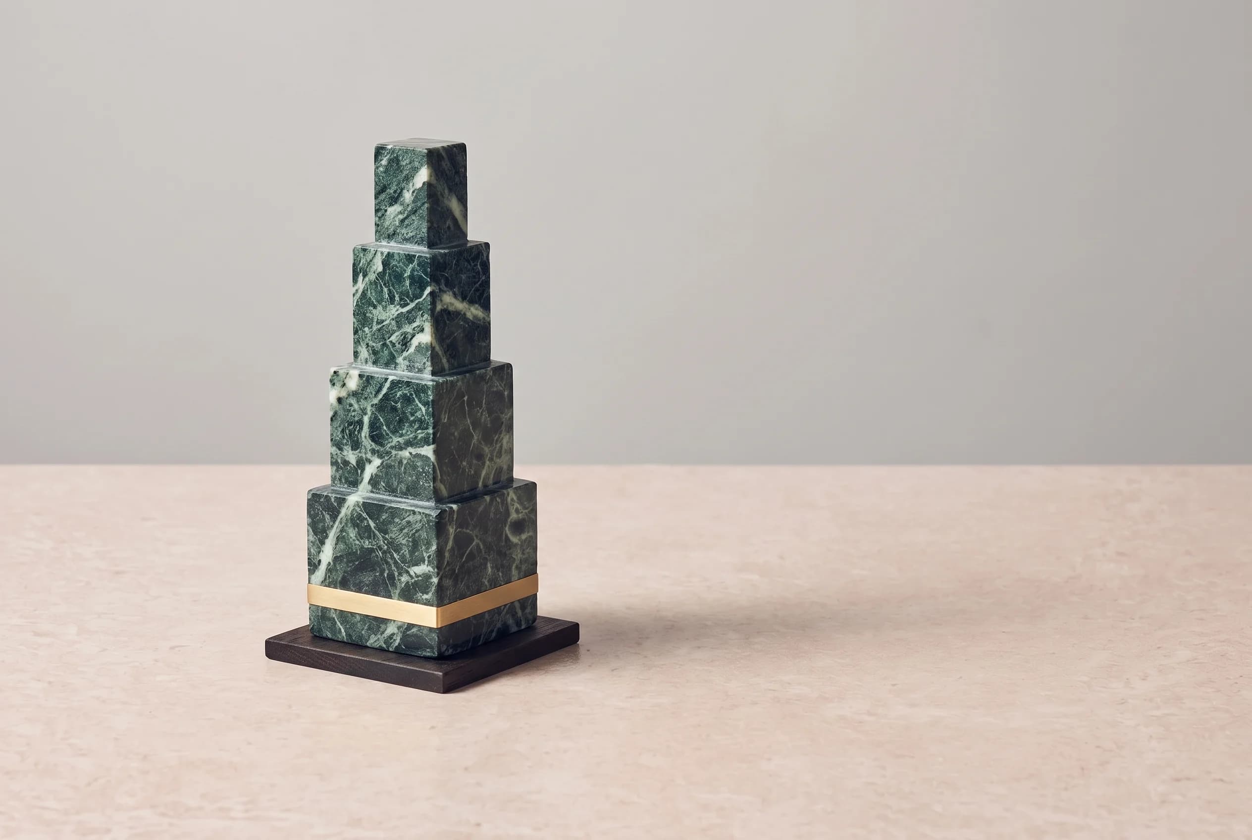 A tall, stepped obelisk of four diminishing marble tiers on a thin brass-edged oak base, standing on a pale stone surface against a grey wall.