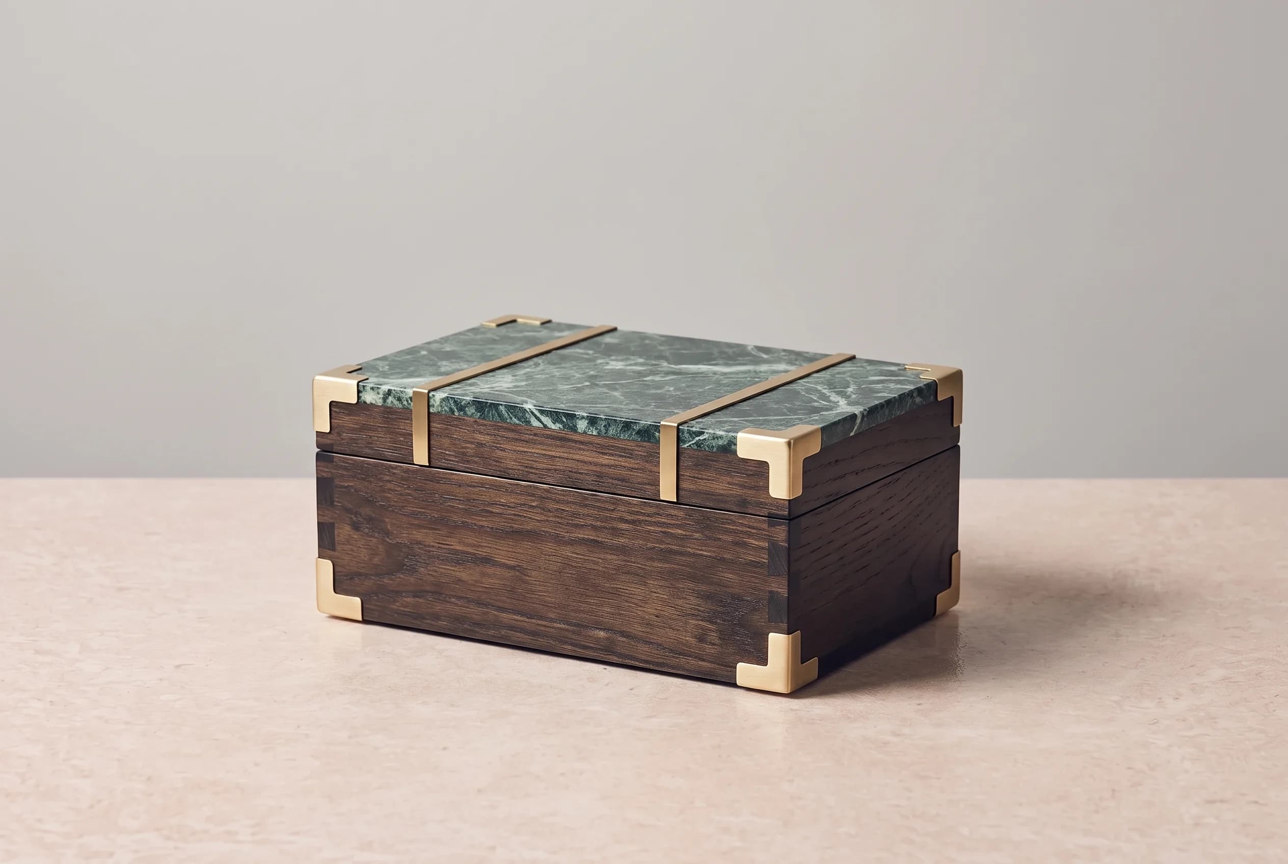 A still life of a rectangular travel trunk in dark smoked oak and Verde Alpi marble with brushed brass straps, standing on a beige stone surface against a grey wall.